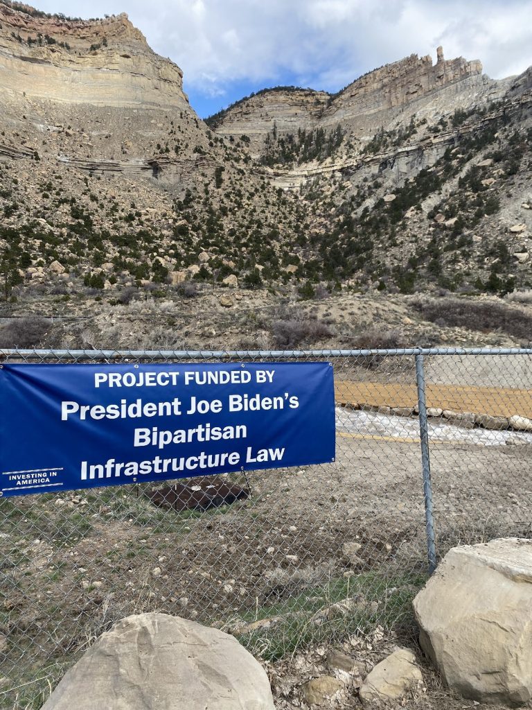 A fence in front of a free-flowing river in a valley with a blue banner that reads “Project Funded by President Joe Biden’s Bipartisan Infrastructure Law”.