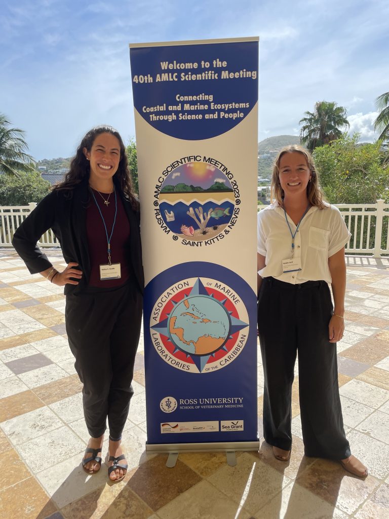 Natalie and Kalina pose for a photo in front of AMLC conference poster in St. Kitts.