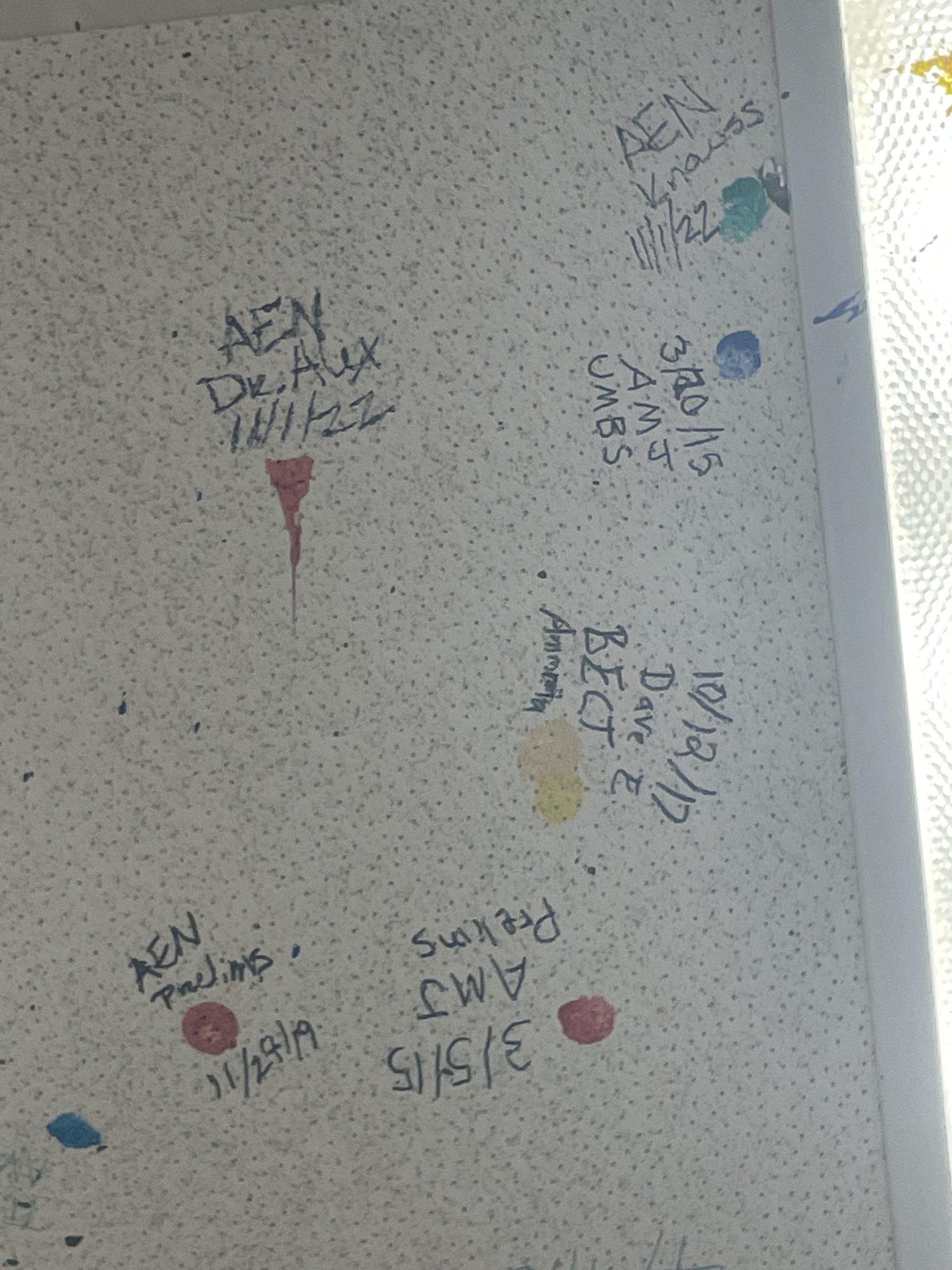 A close up picture of a ceiling tile with red, yellow, and green paint spots. Beside each paint spot are initials, a date, and the accomplishment being celebrated.