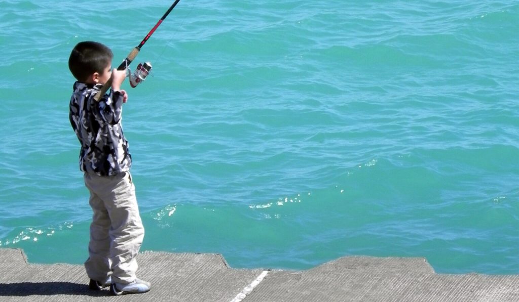 A child holds a fishing rod while standing on a pier near water.
