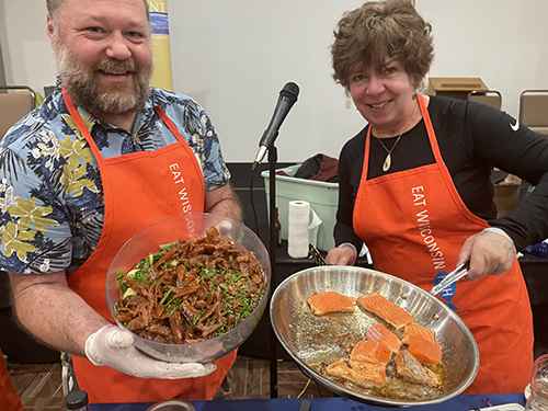 A man and a woman in aprons hold and show two fish-focused dishes.