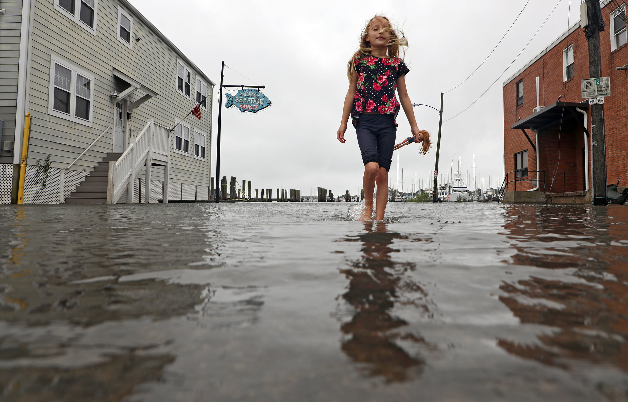 A young girl walks barefoot through a flooded street in front of a seafood market, holding a doll. The street and surrounding buildings are partially submerged, showcasing the effects of coastal flooding in a small town.