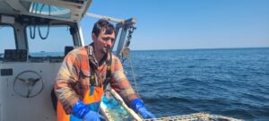 A person wear rubber gloves and coveralls holds a lobster cage over the side of a boat