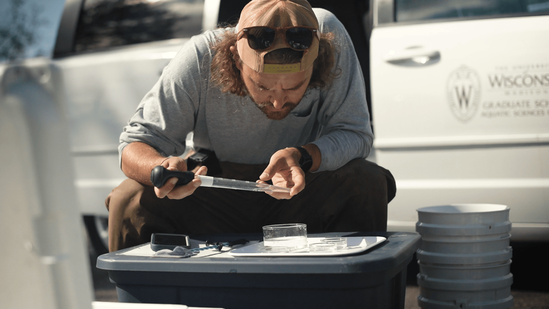 A person sits in front of a van and uses a pipette to examine a water sample sitting on a tray with other equipment.