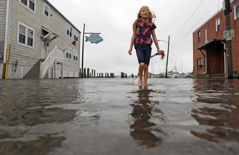 A young girl walks barefoot through a flooded street in front of a seafood market, holding a doll. The street and surrounding buildings are partially submerged, showcasing the effects of coastal flooding in a small town.